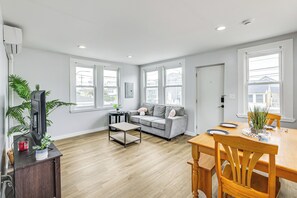 Sala de estar com sofá, mesa de centro e mesa de jantar com cadeiras. Há janelas, uma porta e uma televisão sobre um suporte.