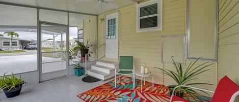 A covered porch with a ceiling fan, a red chair, a green chair, a small table with candles, and potted plants.