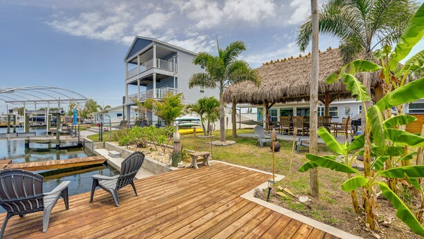 Um deck de madeira com cadeiras, uma piscina, um edifício com varanda e uma estrutura com telhado de palha.