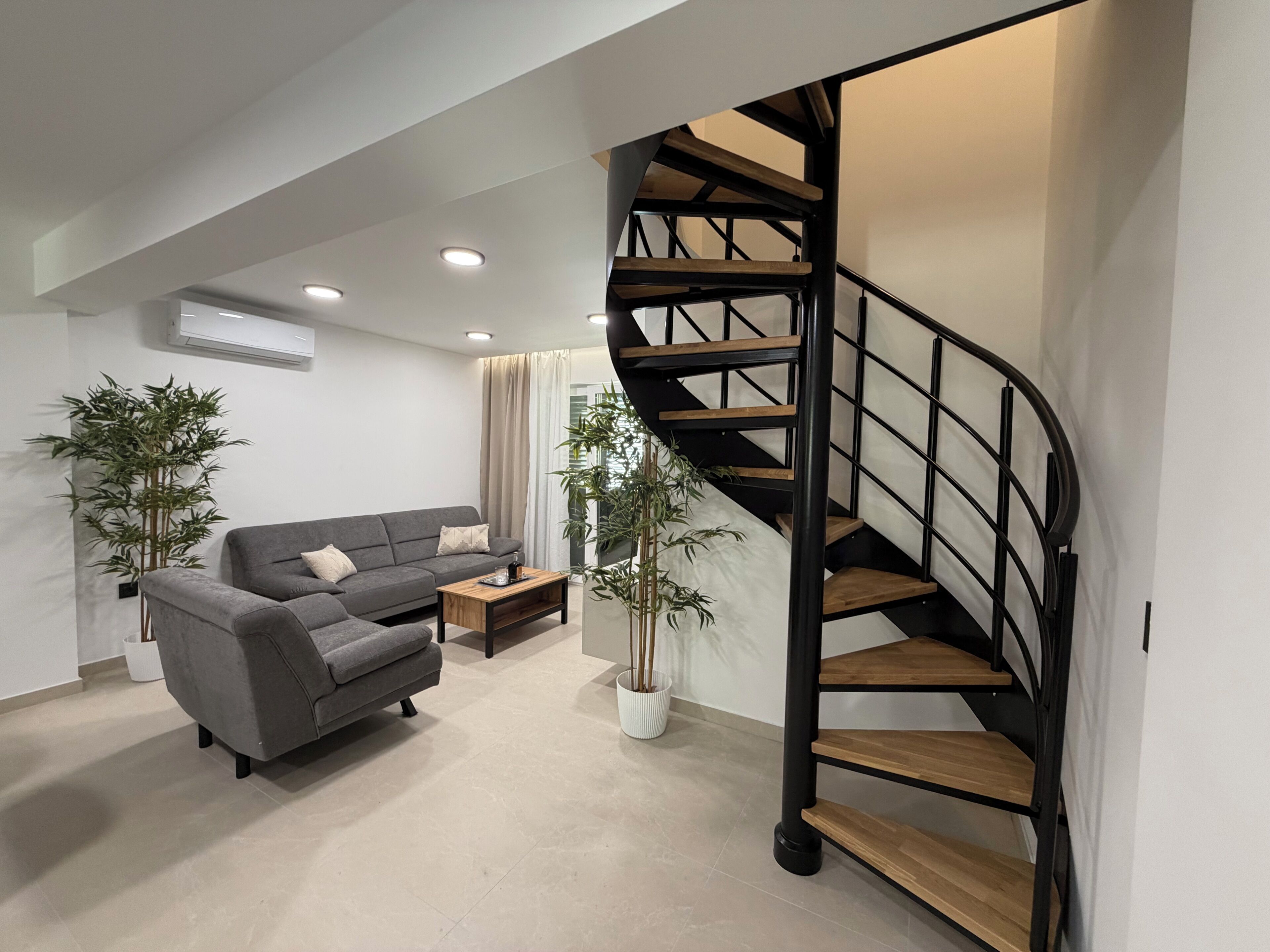 Ein modernes Wohnzimmer mit einem grauen Sofa, einem Holztisch und einer Wendeltreppe.