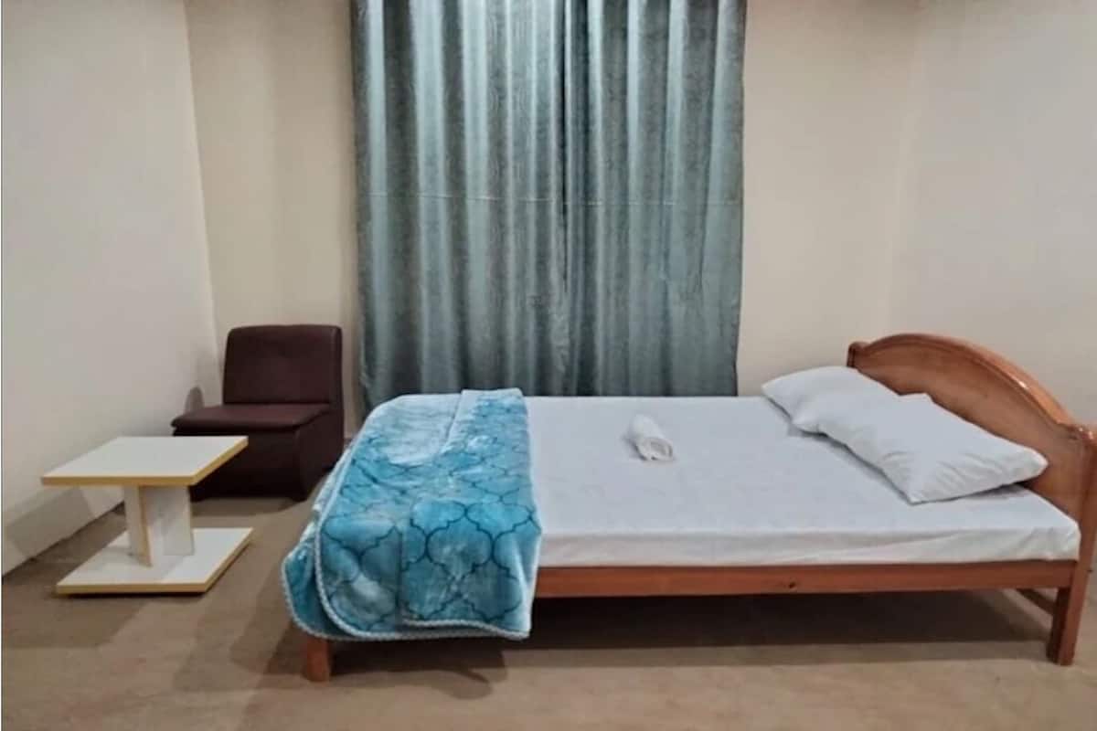 Una cama individual con cabecera de madera, una mesita de noche pequeña, una silla y una ventana con cortinas.