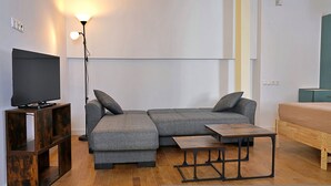 Un salon moderne avec un canapé d’angle, un téléviseur posé sur un meuble en bois, une lampe sur pied et une petite table en bois.