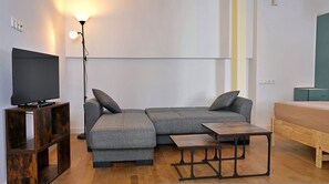 A modern living room with a sectional sofa, a TV on a wooden stand, a floor lamp, and a small wooden table. - Gefyra Studios Filoti Naxos (Naxos)