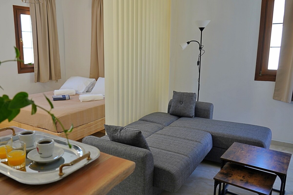 A room with a bed, a grey sofa, a small table with a tray of breakfast, a floor lamp, and a window with curtains.