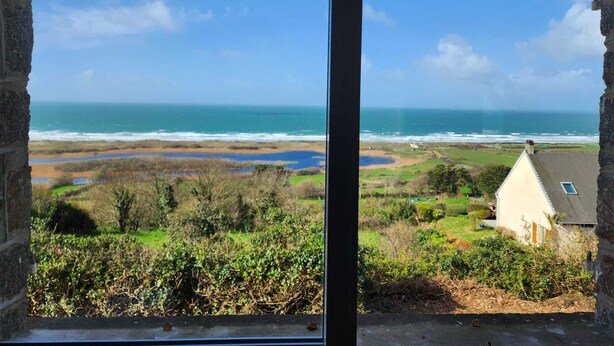 A coastal view through a window with a stone wall, a grassy area, and a building with a sloped roof. - Bernardlhermite (La Hague)