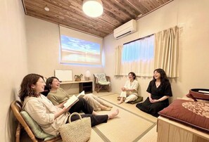 Una habitación con suelo de tatami, pantalla de proyector, ventilador de techo y ventana con cortinas.