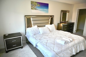 A neatly made bed with white bedding, a nightstand with drawers, a dresser, and a painting of a beach scene on the wall. - Elegant 2-Bedroom Luxury Retreat with 2 Sofa Beds, Close to Niagara Falls (Thorold)