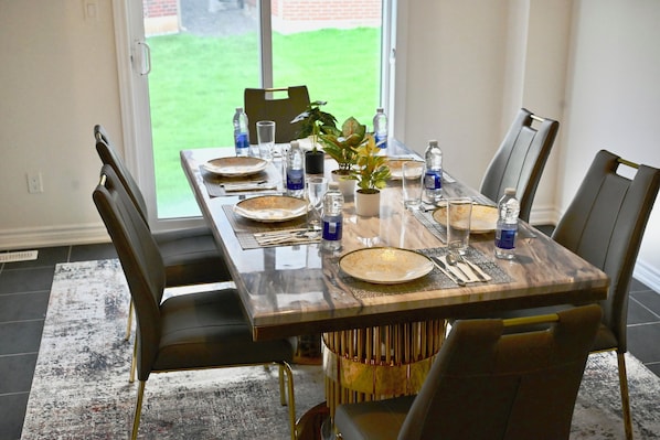 A dining table set with plates, cutlery, and water bottles, surrounded by chairs, with a view of a green lawn through a window.