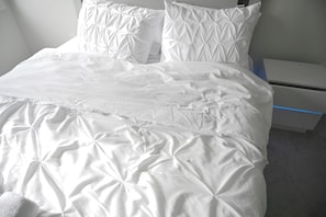 A neatly made bed with a white duvet and pillows, a bedside table with a charging device, and a grey carpeted floor.