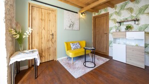 Une pièce avec un plancher en bois, un canapé jaune, une petite table sur laquelle est posé un vase de fleurs, et un coin cuisine avec un meuble blanc et une étagère pour les plantes.