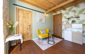 A room with wooden floors, a yellow sofa, a small table with a vase of flowers, and a kitchen area with a white cabinet and a plant shelf. - Awesome home in Marina (Marina)