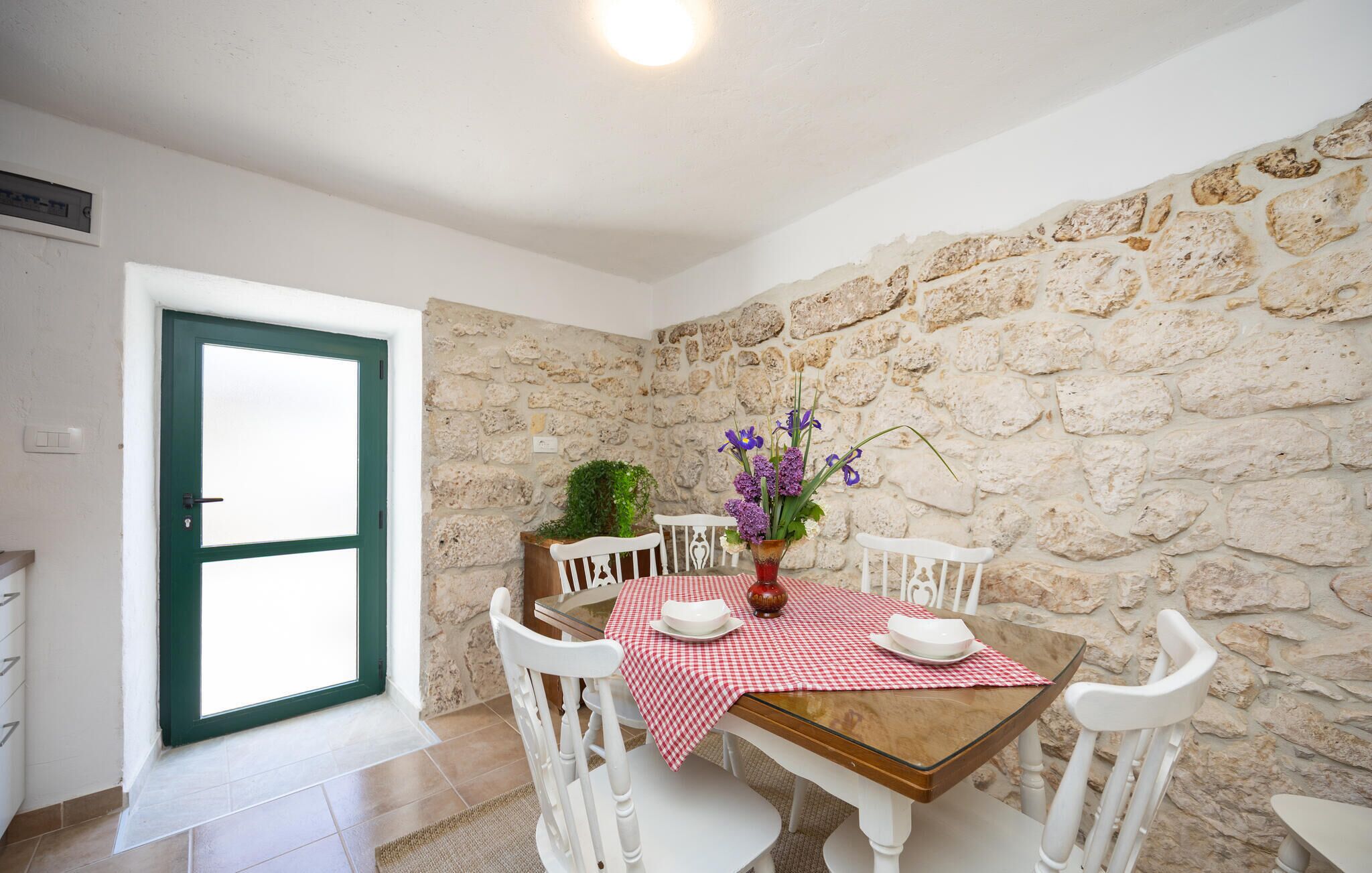 Un comedor con pared de piedra, una mesa de madera, sillas blancas y mantel a cuadros rojo y blanco.