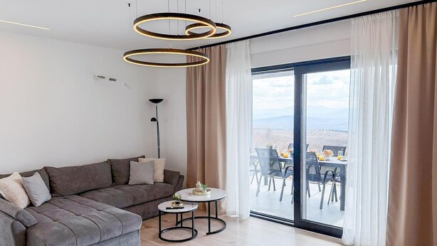 Ett modernt vardagsrum med en grå soffa, ett litet runt bord med en vas och en stor skjutdörr i glas som leder ut till en matplats utomhus.