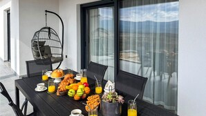 En frukostuppsättning med croissanter, frukt och drycker på ett bord med stolar, nära ett fönster med utsikt över en byggnad.
