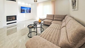 Ein modernes Wohnzimmer mit einem großen beigen Sofa, zwei kleinen runden Tischen, einem Flachbildfernseher und einem Kamin.