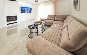 Ein modernes Wohnzimmer mit einem großen beigen Sofa, zwei kleinen runden Tischen, einem Flachbildfernseher und einem Kamin.