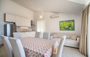 A compact living space with a kitchenette, a dining table with patterned tablecloth, a sofa with patterned cushions, and a painting on the wall. - Cozy apartment in Bast with swimming pool (Bast)