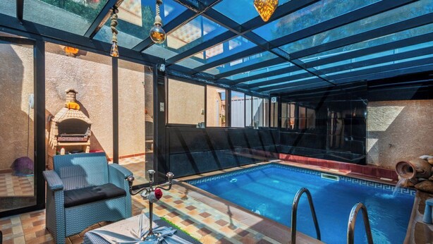 En swimmingpool med glasvægge, en murstensovn, fletningsmøbler og flisegulv.