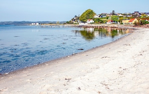 Uma praia costeira com águas claras, areal arenoso e casas ao fundo.