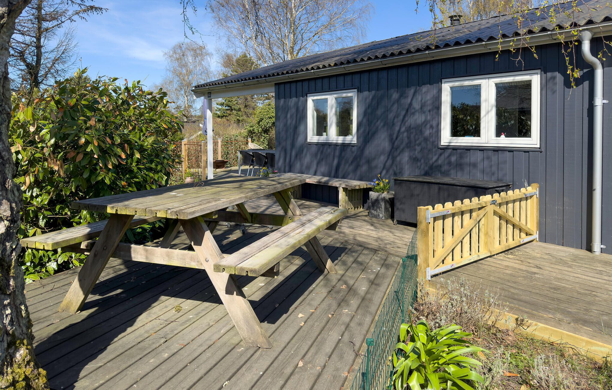 Een houten terras met een picknicktafel, een tuinhuisje met twee ramen, en een houten poort.