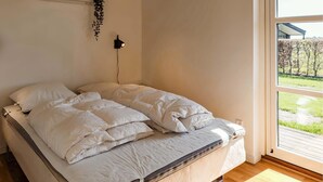 Un lit avec une literie blanche, une lampe fixée au mur et une vue sur un jardin par la fenêtre.