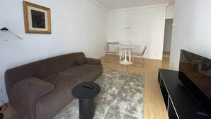 Een moderne woonkamer met een bank, een klein rond tafeltje, een televisie en een eethoek met een tafel en stoelen.