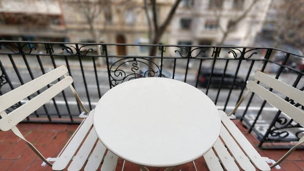 Een witte buitentafel met stoelen op een met bakstenen bestraat gebied, met een metalen hekwerk, uitkijkend over een straat met gebouwen en geparkeerde auto's.