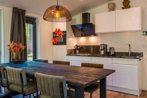 A modern kitchen with a dining area, featuring a large wooden table, cushioned chairs, a hanging light fixture, and a Buddha statue. - De Wiltzangh - Zandborg Lodge Hottub | 6p (Ruinen)