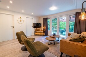 Ein modernes Wohnzimmer mit einer Couch, Sesseln, einem Couchtisch, einem Fernseher und einer Schiebetür zum Außenbereich.