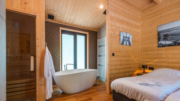 Interior de una cabaña de madera con una bañera grande, una cama y una ventana con vistas.