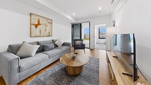 Uma sala de estar moderna com um sofá cinza, uma mesa de centro de madeira, uma poltrona preta e uma TV sobre um suporte de madeira.