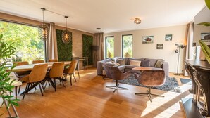 A modern living room with a wooden floor, a sectional sofa, a dining area with chairs, and a large window with a view of greenery. - Vakantiepark De Wiltzangh - De Havezate Wellness (Ruinen)