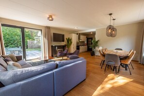 A modern living room with a large sofa, dining table, and chairs, a kitchen in the background, and a large window with a view of greenery.