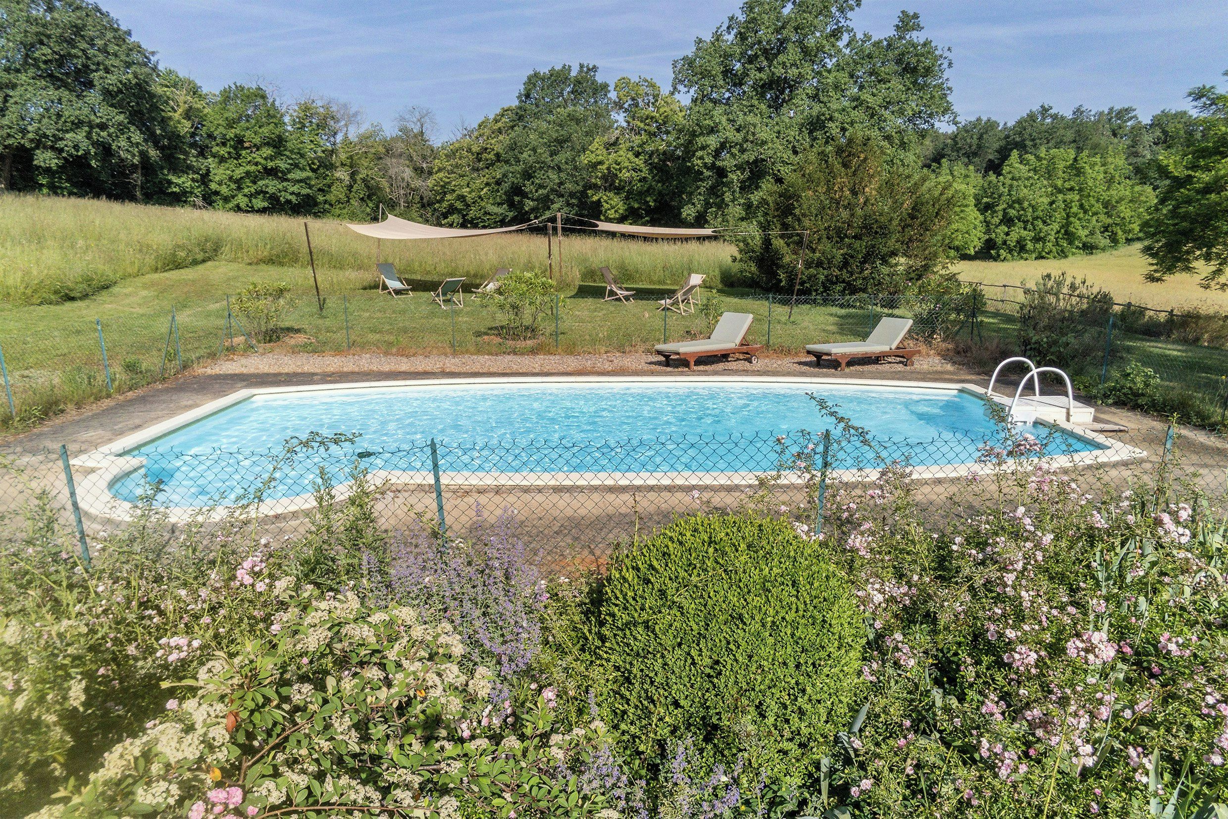 A rectangular swimming pool with a chain-link fence, surrounded by greenery and a few lounge chairs.