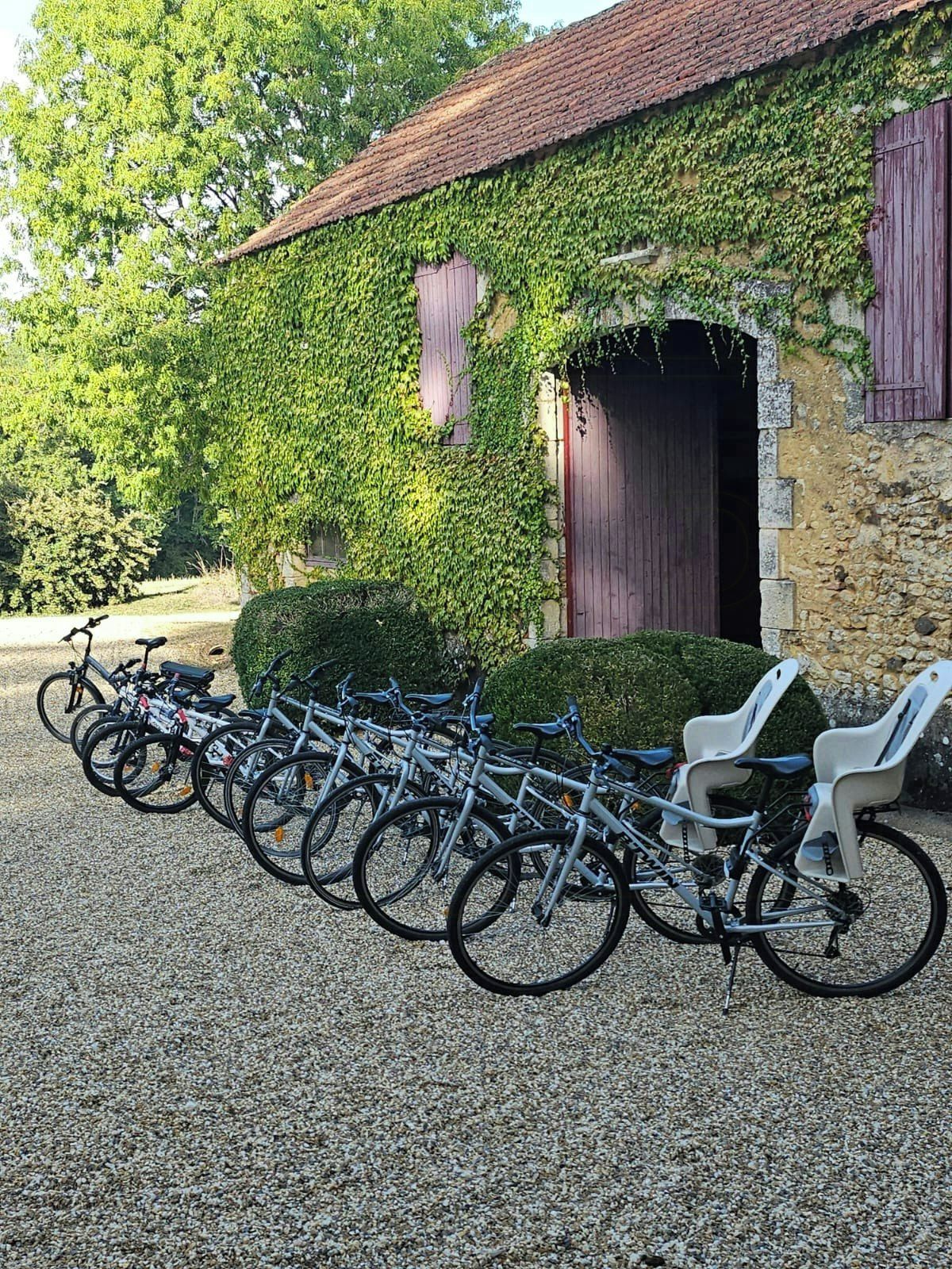 A row of bicycles parked in front of a stone building with a tiled roof, surrounded by greenery.