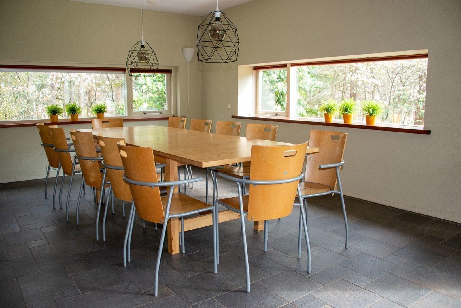 Un salón de conferencias con una mesa y sillas de madera, ventanas amplias con plantas y lámparas geométricas colgantes.