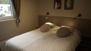 Una cama bien hecha con cubrecama acolchada, dos almohadas decorativas, una lámpara de noche y una ventana con cortinas.
