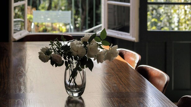 Un tavolo da pranzo con un vaso di fiori, lampade a sospensione e vista attraverso una porta aperta su un balcone con ringhiera.