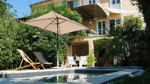 Area piscina con lettini e ombrellone, immersa nel verde e affiancata da un edificio con balconi.
