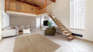 Een ruime woonkamer met een hoge nok, een houten trap en een groot raam.