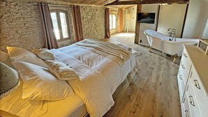 Une chambre à coucher avec un grand lit, un plafond en bois, un mur en pierre, une fenêtre et une salle de bain avec une baignoire.