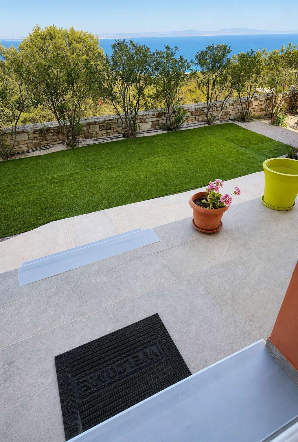 A patio with a view of the sea, a green lawn, and a welcome mat.