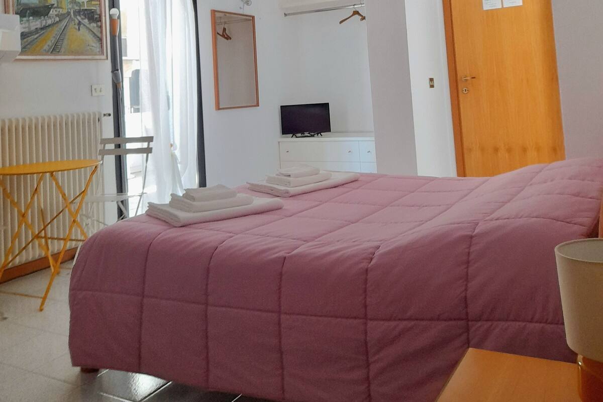 A hotel room with a bed, a TV, a wooden door, and a painting on the wall.