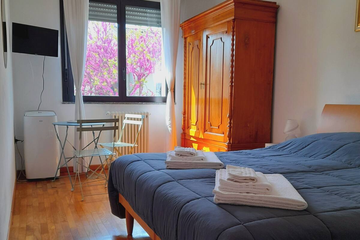 A bedroom with a bed, a wooden wardrobe, a desk with a chair, a radiator, and a window with curtains.