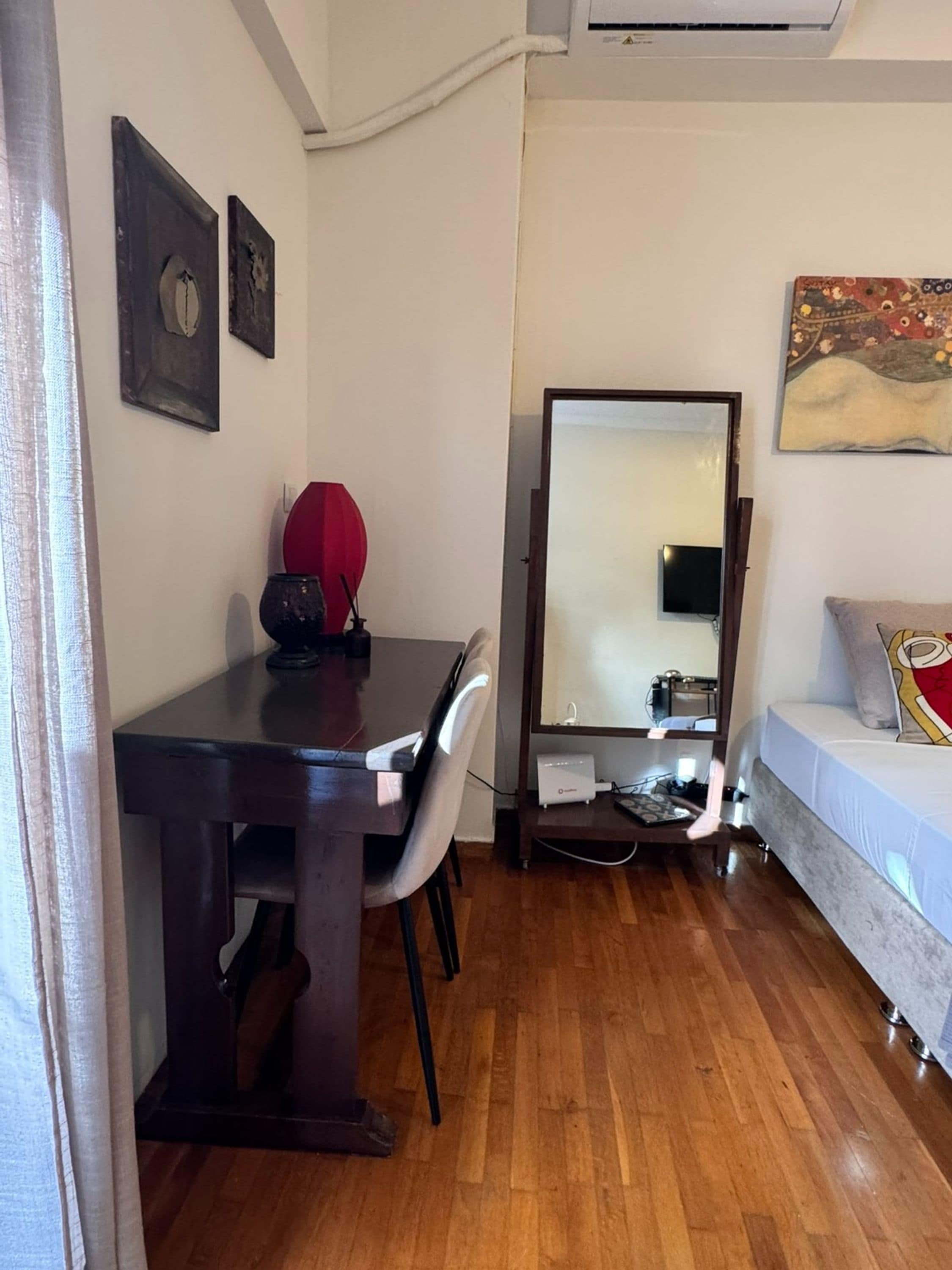 A compact room with a wooden floor, a dining table with chairs, a sofa, a large mirror, and a painting on the wall.