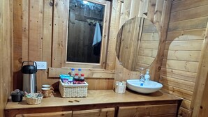 Une salle de bain de style rustique, avec des murs en bois, une fenêtre, un lavabo, un miroir, un porte-serviettes, un panier contenant des articles de toilette et un thermos.