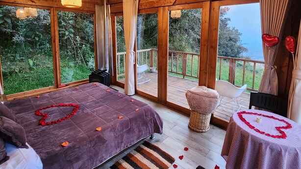 Une cabane en bois avec un grand lit, une petite table recouverte d’une nappe en forme de cœur et un balcon offrant une vue sur la verdure.