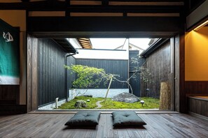 A traditional Japanese room with tatami flooring, a low wooden ceiling, and a view of a garden with rocks, moss, and a tree.