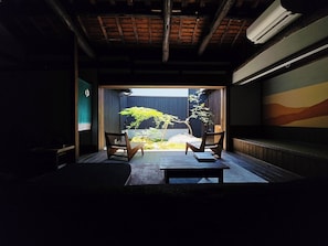 Une pièce de style japonais traditionnel, avec un sol recouvert de tatamis, une table basse, deux chaises et une vue sur un jardin, accessible par une porte coulissante.