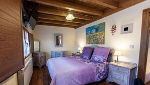 Une chambre avec un plafond en bois, un lit recouvert d’une couverture violette, une table de chevet avec une lampe, une commode et un tableau accroché au mur.